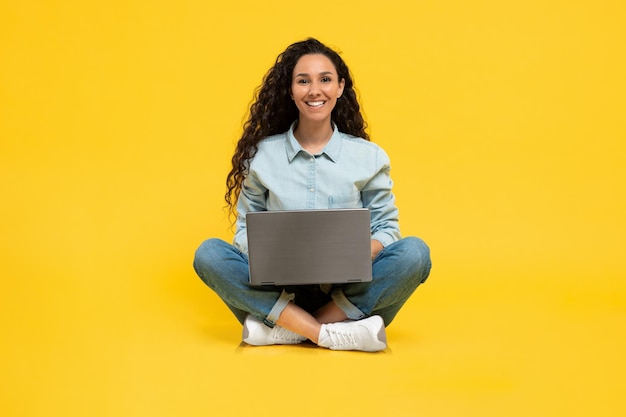 Foto mujer joven feliz que usa una computadora portátil para trabajar o estudiar en línea sentada con las piernas cruzadas en el fondo del estudio amarillo