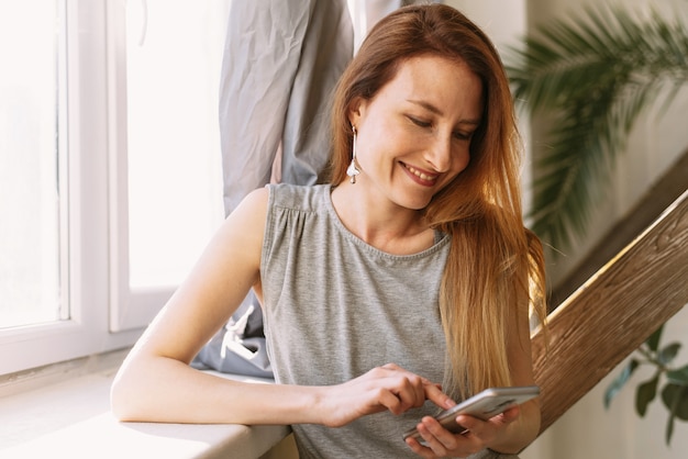 Mujer joven feliz que usa el teléfono móvil en casa con la computadora portátil