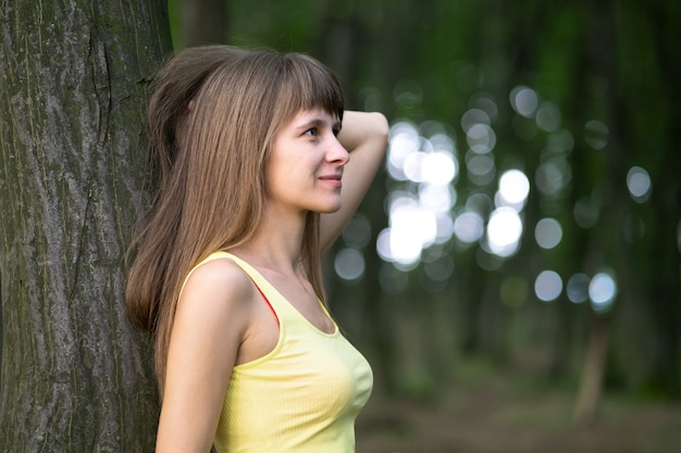Mujer joven feliz relajante mientras se inclina a un gran tronco de árbol en el parque de verano.