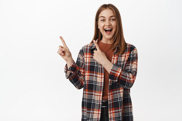 Mujer joven feliz riendo y sonriendo, señalando la esquina superior izquierda, invitando a ver los descuentos promocionales en la tienda, mostrando el logotipo publicitario, fondo blanco.