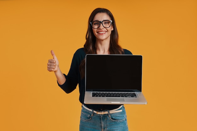 Mujer joven feliz en ropa casual elegante que señala el espacio de la copia en la computadora portátil