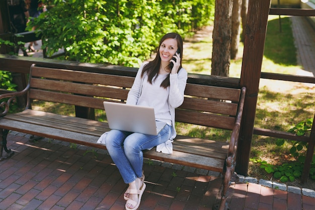 Mujer joven feliz en ropa casual ligera hablando por teléfono móvil. Mujer sentada en un banco trabajando en un moderno ordenador portátil en la calle al aire libre en la naturaleza. Oficina móvil. Concepto de negocio autónomo.