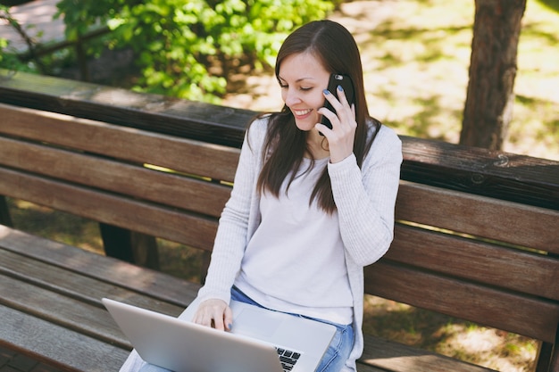 Mujer joven feliz en ropa casual ligera hablando por teléfono móvil. Mujer sentada en un banco trabajando en un moderno ordenador portátil en la calle al aire libre en la naturaleza. Oficina móvil. Concepto de negocio autónomo.