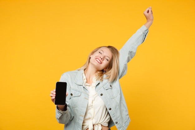 Mujer joven feliz con ropa casual de mezclilla manteniendo los ojos cerrados, levantando la mano para sostener el teléfono móvil con una pantalla vacía en blanco aislada en un fondo amarillo anaranjado. Concepto de estilo de vida de las personas. Simulacros de espacio de copia.