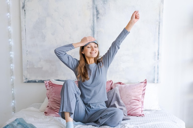 Mujer joven feliz sentada en una cama cómoda con los brazos extendidos después de despertarse