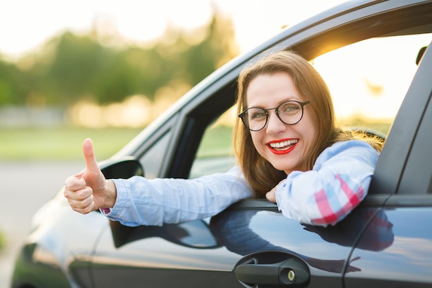 Mujer joven feliz sentada en un coche con el pulgar hacia arriba concepto de comprar un coche usado o un coche de alquiler