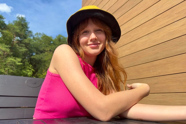 Mujer joven feliz sentada en la terraza en la mesa al aire libre en el día de verano en sombrero de Panamá y sonrisa mirando a la cámara