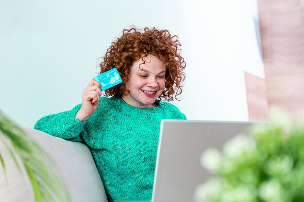 Mujer joven feliz en el sofá comprando en línea con tarjeta de débito Niña hermosa usando computadora portátil para comprar en línea en casa salió para encontrar un artículo en venta