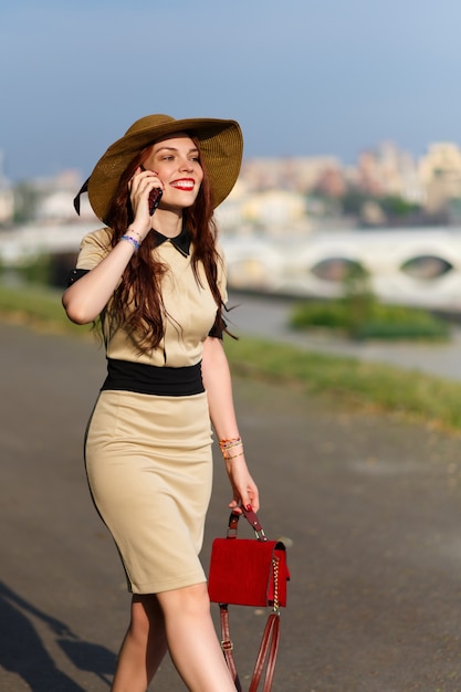 Una mujer joven feliz con un sombrero de ala ancha está caminando en el parque en el verano con una bolsa y hablando por teléfono