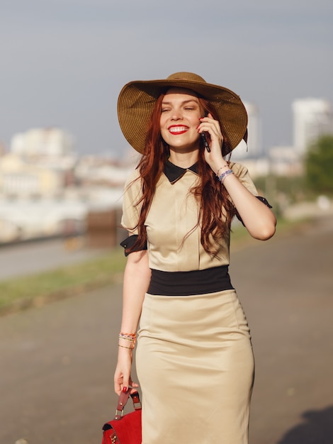 Una mujer joven feliz con un sombrero de ala ancha está caminando en el parque en el verano con una bolsa y hablando por teléfono