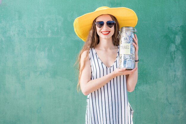 Mujer joven y feliz con sombrero amarillo sosteniendo un frasco lleno de dinero para viajar en el fondo verde. Ahorro de dinero por concepto de vacaciones de verano
