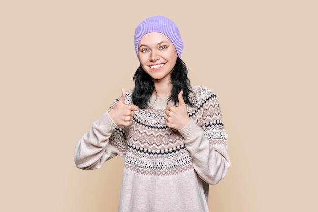 Foto mujer joven feliz con sombrero de suéter de invierno mostrando los pulgares hacia arriba sobre fondo marrón