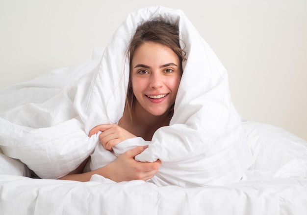 Mujer joven feliz sonriente cubierta con una manta caliente en la cama en casa por la mañana