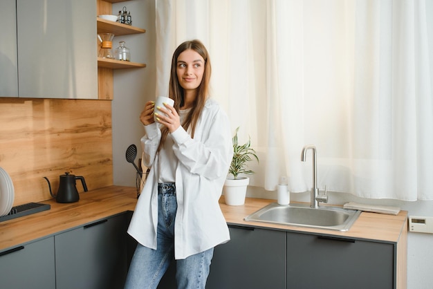 Mujer joven feliz sosteniendo una taza de té mientras está de pie en la cocina en casa por la mañana Concepto de estilo de vida saludable