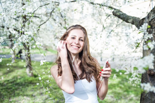 Mujer joven feliz sosteniendo el teléfono y escuchando música o podcast a través de auriculares en una camiseta blanca bajo los cerezos en flor Sonriendo mirando a la cámara