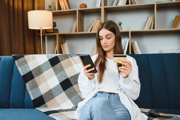 Mujer joven feliz sosteniendo teléfono y tarjeta de crédito sentado en el sofá Compras en línea