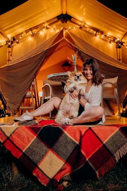 Mujer joven feliz con su perro Welsh Corgi Pembroke relajándose en glamping en la noche de verano cerca de una acogedora hoguera Tienda de campaña de lujo para recreación al aire libre y recreación Concepto de estilo de vida