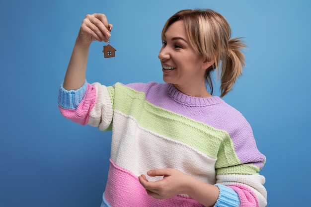 Mujer joven feliz en suéter casual sosteniendo llavero de su nuevo apartamento sobre fondo azul con