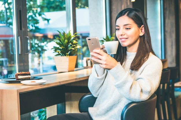 Mujer joven feliz con una taza de café sentado junto a una ventana en la cafetería hablando por teléfono