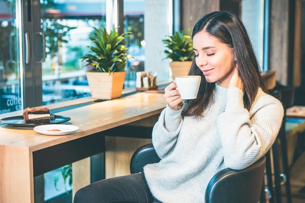 Mujer joven feliz con una taza de café sentado junto a una ventana en la cafetería con los ojos cerrados