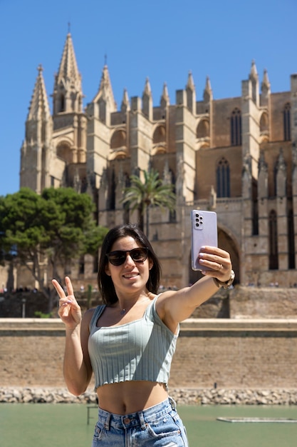 Mujer joven feliz tomando una foto con su teléfono móvil frente a la catedral de Palma de Mallorca en España en un día soleado de verano