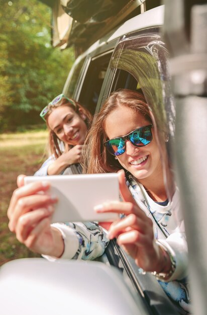 Mujer joven feliz tomando fotos con un teléfono inteligente a su amiga sentada detrás de la ventana del coche en una aventura de viaje por carretera. Amistad femenina y concepto de tiempo libre.