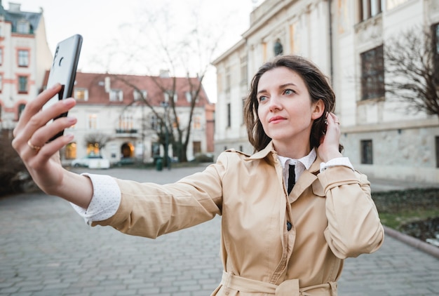 Mujer joven feliz tomando selfie en la calle de la ciudad. Chica turista feliz caminando al aire libre. Retrato de moda de primavera de mujer bonita morena posando en el casco antiguo.