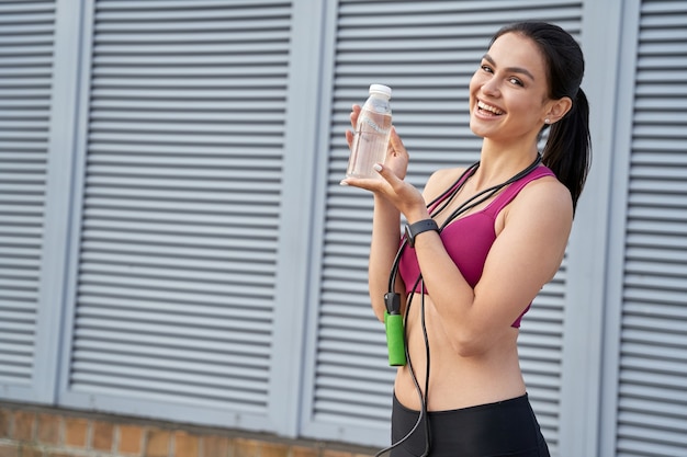 Mujer joven feliz en un top deportivo y polainas que demuestran una botella de agua. Concepto de deporte. Copia espacio