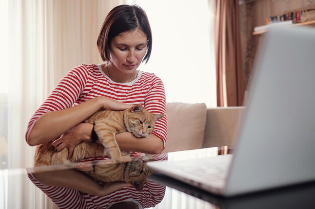 Mujer joven feliz trabaja en casa con una computadora portátil y un gato de jengibre