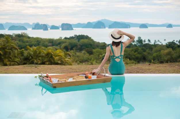 Mujer joven feliz en traje de baño verde con bandeja de desayuno flotante en la piscina de lujo hotel chica con sombrero disfrutar en un resort tropical Relajante Vacaciones de verano exóticas viajes y vacaciones tropicales