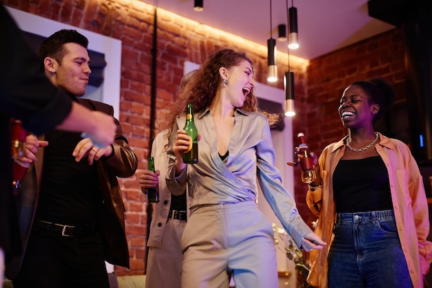 Mujer joven feliz en traje gris con una botella de cerveza bailando entre amigos disfrutando de casa pa