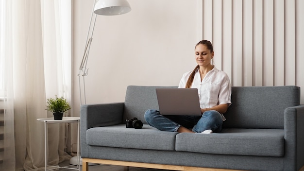 Mujer joven feliz usando la computadora portátil en casa. En el sofá junto a la mujer hay una cámara. Fotógrafo retoca fotografías en casa.