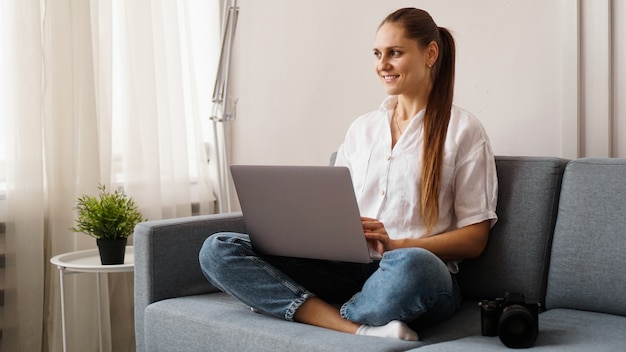 Mujer joven feliz usando la computadora portátil en casa. En el sofá junto a la mujer hay una cámara. Fotógrafo retoca fotografías en casa.