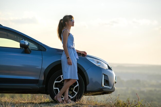 Mujer joven feliz en vestido azul de pie cerca de su vehículo mirando la vista del atardecer de la naturaleza de verano Concepto de viaje y vacaciones