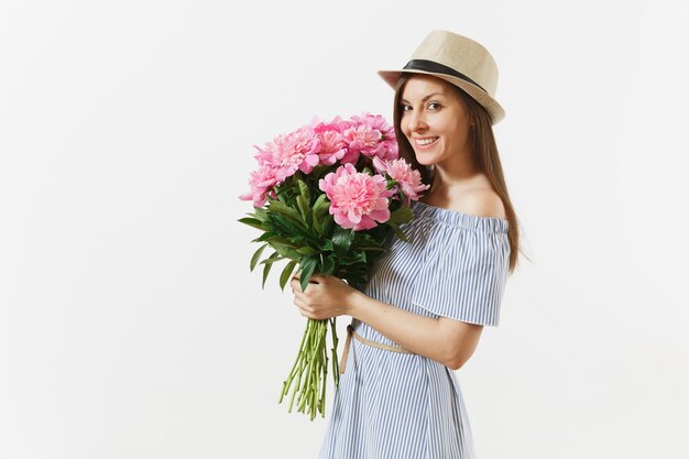 Mujer joven feliz en vestido azul, sombrero con ramo de hermosas flores de peonías rosas aisladas sobre fondo blanco. Día de San Valentín, concepto de vacaciones del Día Internacional de la Mujer. Área de publicidad.