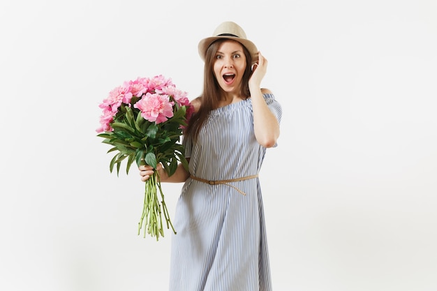 Mujer joven feliz en vestido azul, sombrero con ramo de hermosas flores de peonías rosas aisladas sobre fondo blanco. Día de San Valentín, concepto de vacaciones del Día Internacional de la Mujer. Área de publicidad.