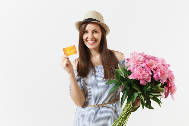 Mujer joven feliz en vestido azul, sombrero con tarjeta de crédito, dinero, ramo de hermosas flores de peonías rosas aisladas sobre fondo blanco. Negocio, entrega, concepto de compra online. Copie el espacio.