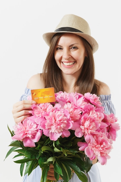 Mujer joven feliz en vestido azul, sombrero con tarjeta de crédito, dinero, ramo de hermosas flores de peonías rosas aisladas sobre fondo blanco. Negocio, entrega, concepto de compra online. Copie el espacio.