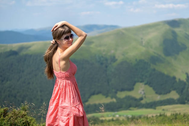 Mujer joven feliz en vestido rojo caminando sobre el campo de hierba en un día ventoso en las montañas de verano.