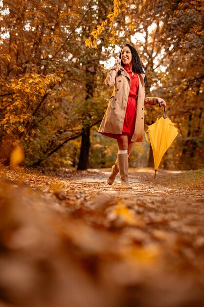 Mujer joven feliz con vestido rojo hablando por teléfono inteligente caminando en el parque soleado de otoño, sosteniendo un paraguas amarillo doblado.