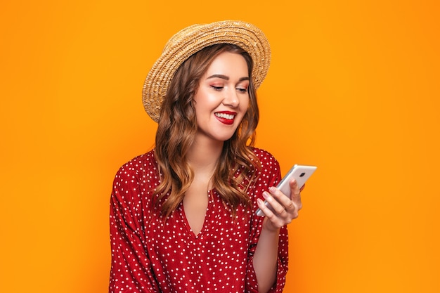 Mujer joven feliz en un vestido rojo del sombrero de paja con el lápiz labial rojo que mira su teléfono móvil y sonrisa aislada en la pared amarilla. Niña lee un mensaje hace compras en línea