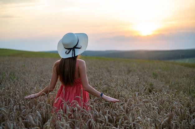 Mujer joven feliz en vestido rojo de verano y sombrero de paja blanco de pie en el campo agrícola amarillo con trigo dorado maduro disfrutando de una cálida noche