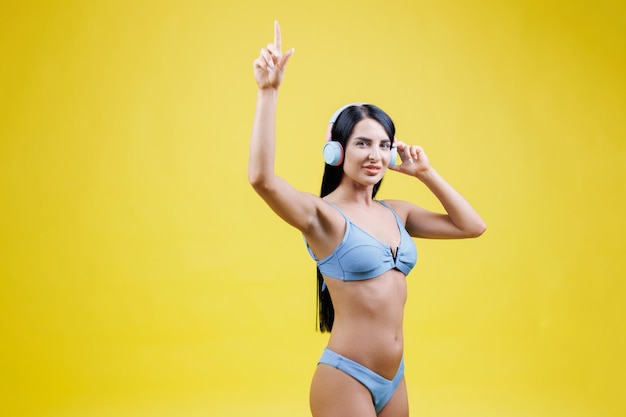 Mujer joven feliz vistiendo traje de baño azul escuchando música en auriculares y bailando cantando y divirtiéndose aislado en el estudio de fondo de pared amarilla Concepto de descanso de verano