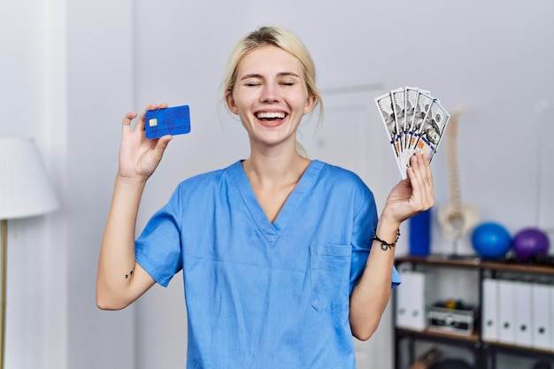 Mujer joven fisioterapeuta con tarjeta de crédito y billetes de dólares sonriendo y riendo a carcajadas porque es una broma divertida y loca