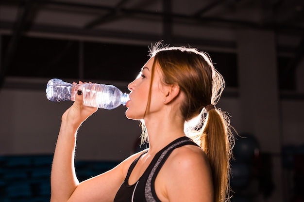 Mujer joven fitness agua potable en el gimnasio. Mujer muscular tomando un descanso después del ejercicio