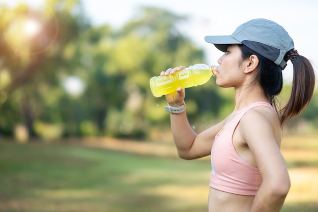 Mujer joven fitness bebiendo agua de energía durante el estiramiento muscular en el parque al aire libre, atleta asiático corriendo y ejercicio por la mañana Concepto de deporte y bienestar