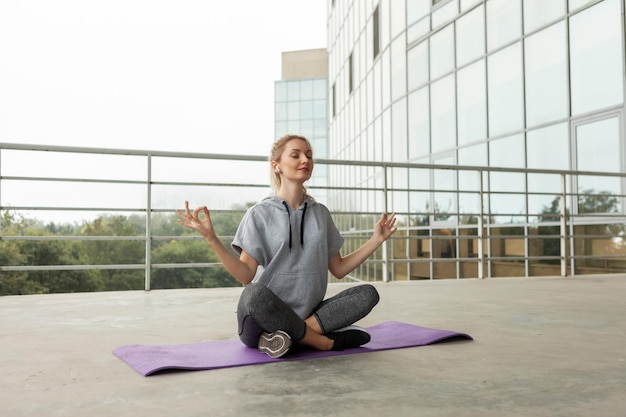 Mujer joven fitness en ropa deportiva practicando yoga, medita en la azotea del edificio, estilo urbano