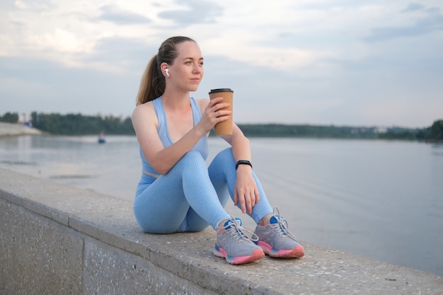 Mujer joven fitness terminar corriendo en la ciudad. disfruta de un café mientras escuchas música en los auriculares. atardecer. estilo de vida saludable. libertad. entrenamiento al aire libre. Foto de alta calidad