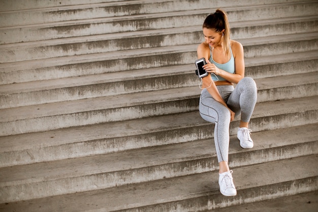 Mujer joven fitness tomando un descanso de correr sentado en los pasos y demandando teléfono móvil en brazalete