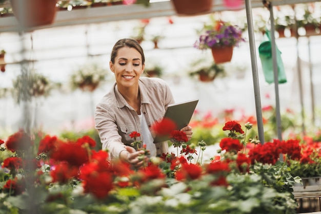 Mujer joven florista usando tableta digital mientras revisa flores en un invernadero.